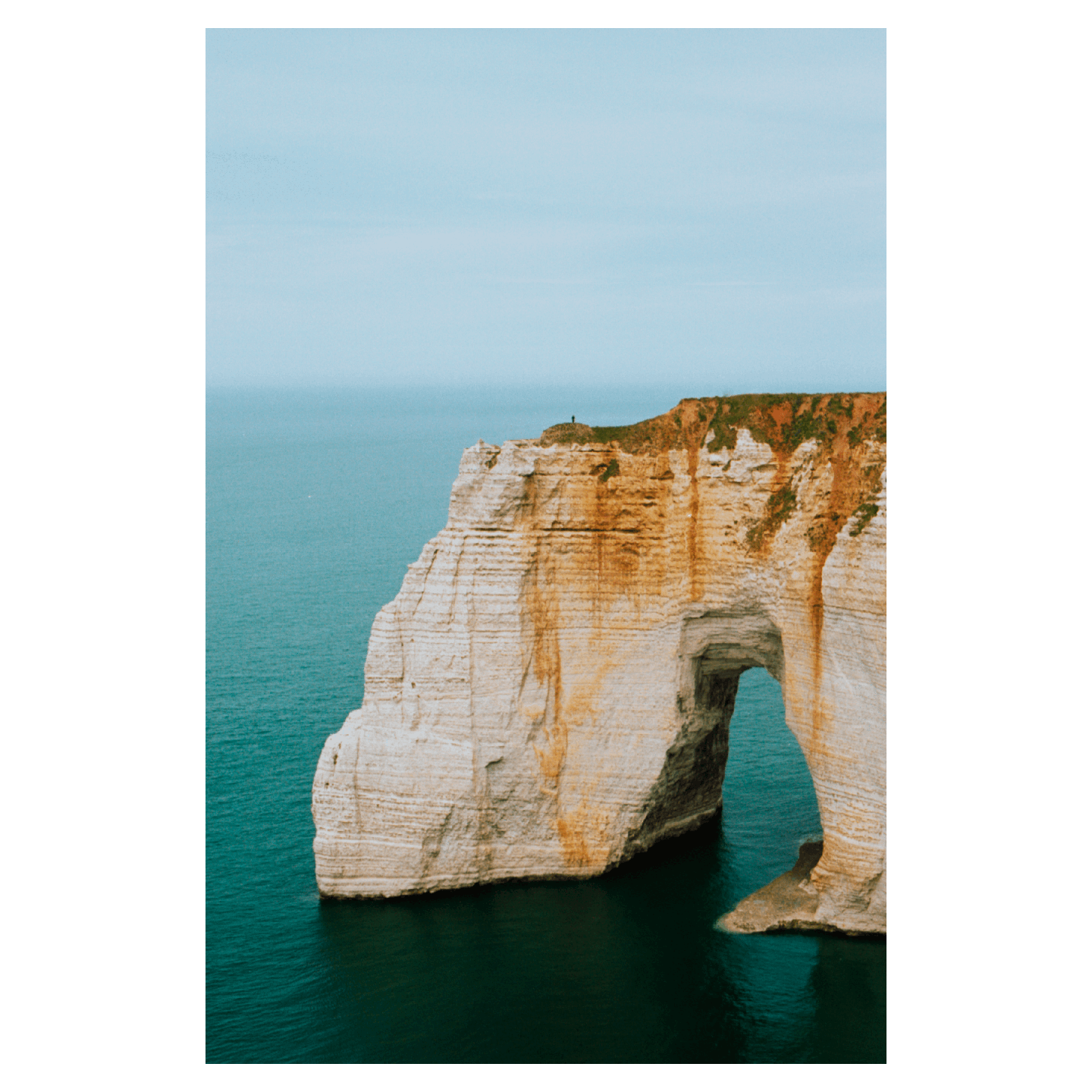 A person stands atop a large cliff arching over the ocean.