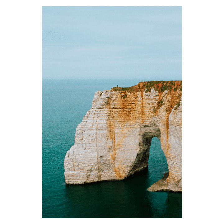 A person stands atop a large cliff arching over the ocean.