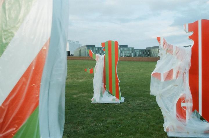 A red and green striped bear statue stands in a grassy field with buildings in the background.