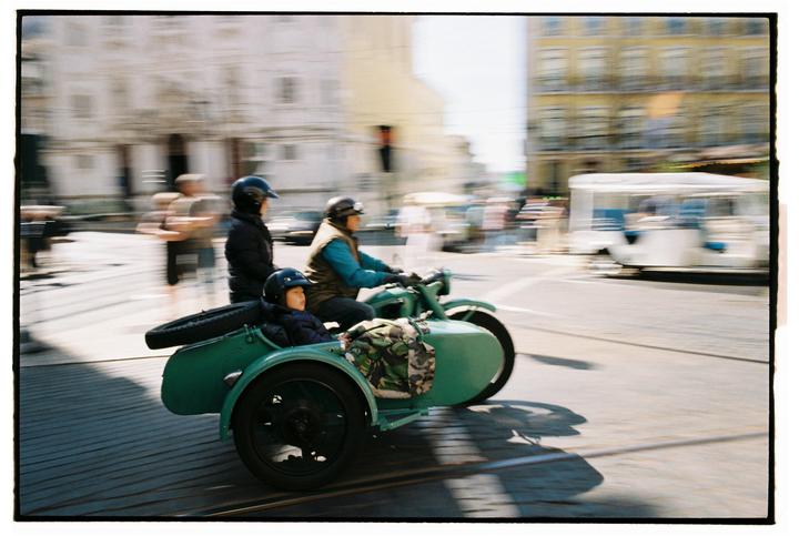 A man drives a green motorcycle with a sidecar carrying a child and another man down a city street.