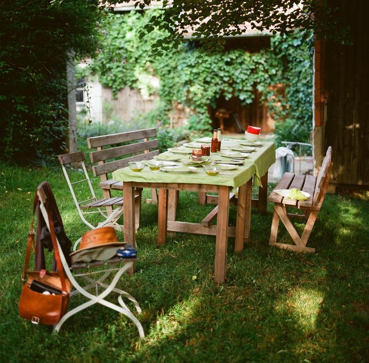 A wooden table set with plates and bowls sits on a grassy lawn surrounded by trees and benches.