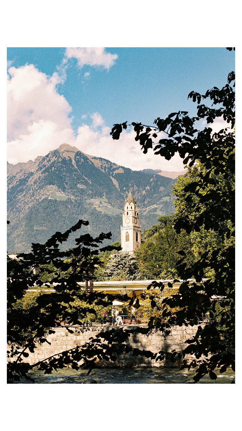A tall clock tower stands among trees with a mountain in the background and a river in the foreground.