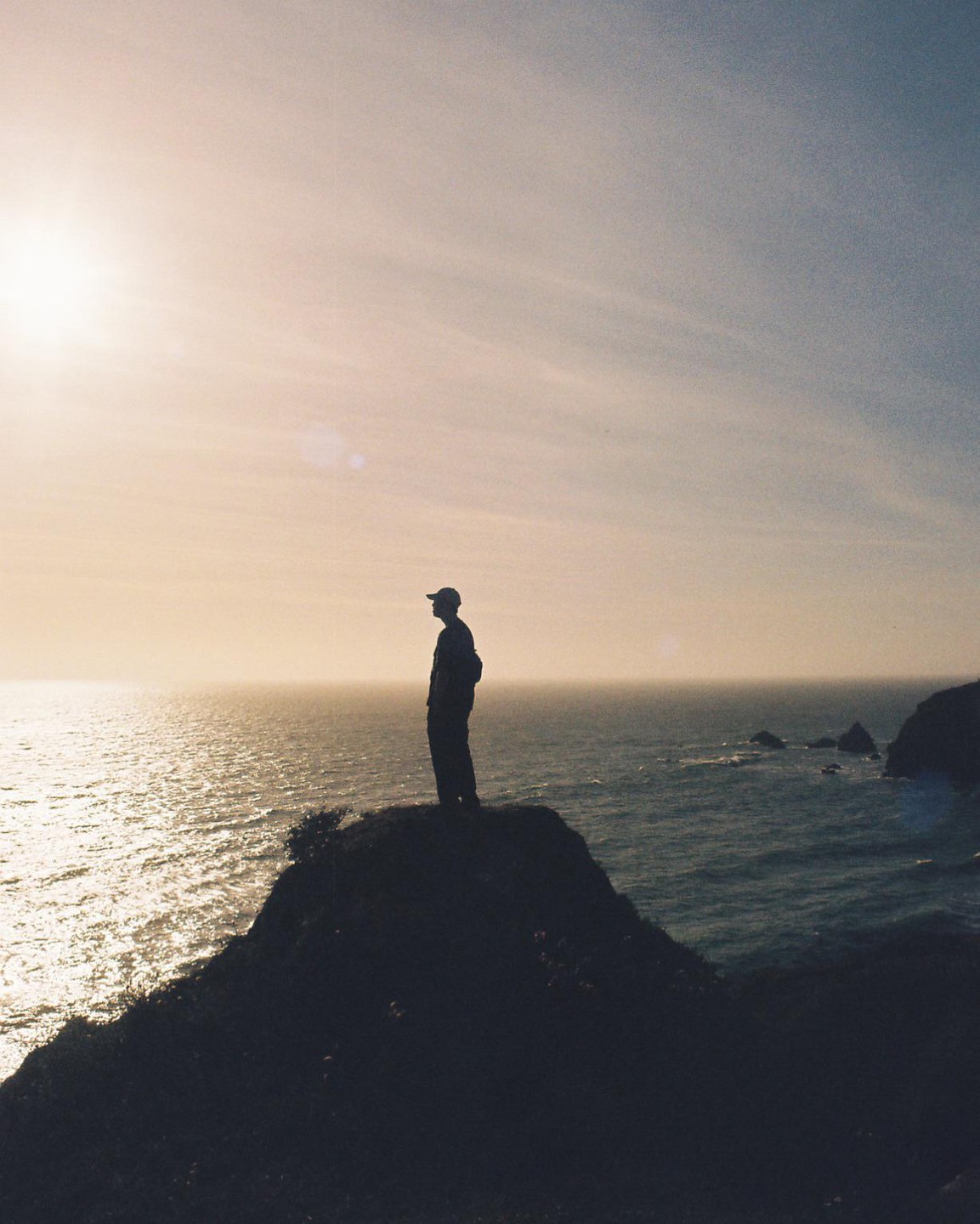 A man stands silhouetted on a cliff overlooking the ocean at sunset.