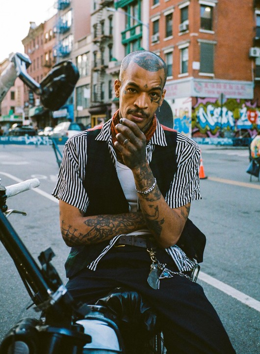A man with tattoos on his face and arms sits on a motorcycle on a city street.