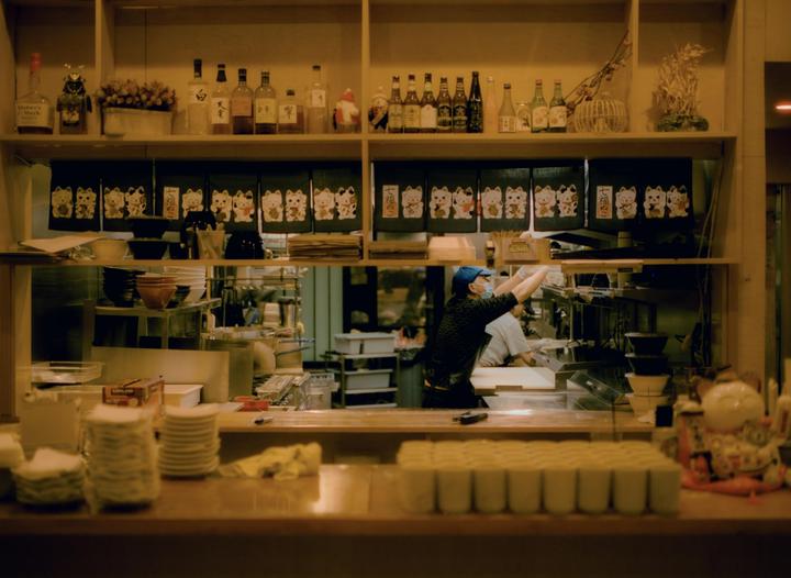 A man wearing a face mask and a blue cap is working in a kitchen with shelves of bottles and cat-patterned banners above him.