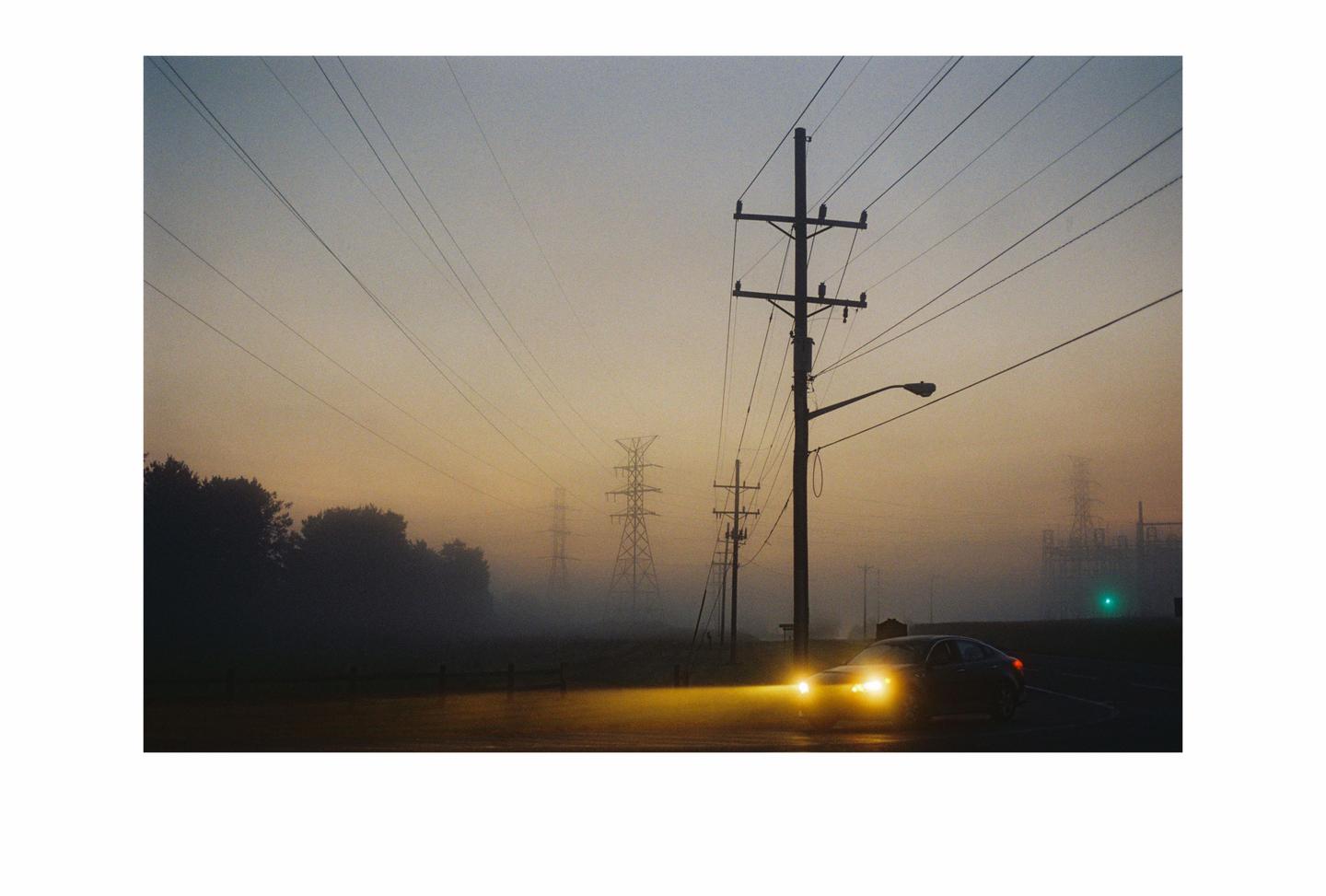 A car with its headlights on drives on a road with power lines and transmission towers in the fog at dawn.