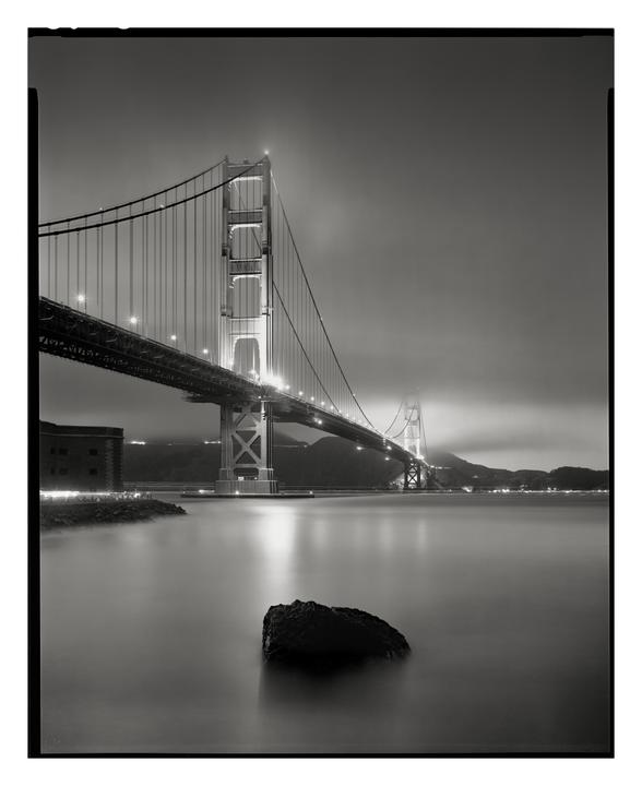 The Golden Gate Bridge is illuminated at night with a large rock in the foreground and a building on the left.