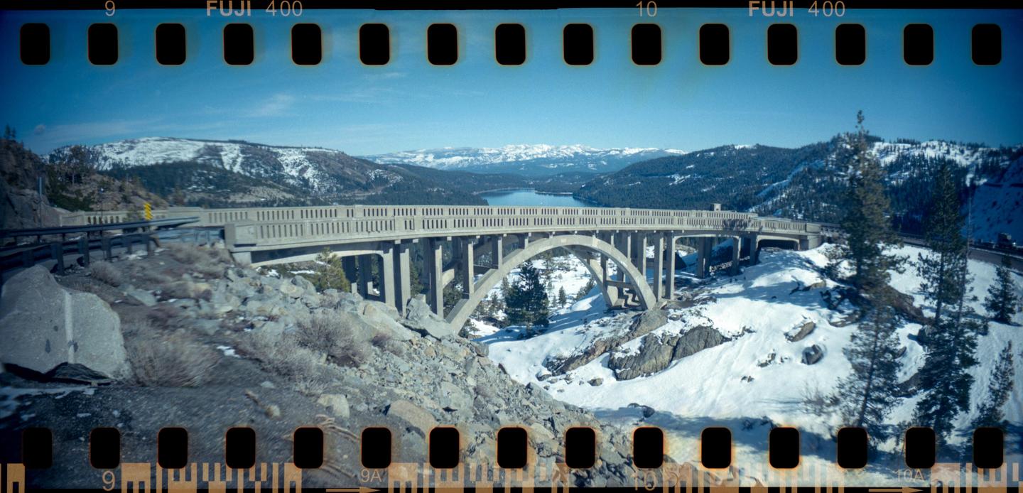 A concrete bridge with arches spans a snowy landscape with a lake and mountains in the distance.