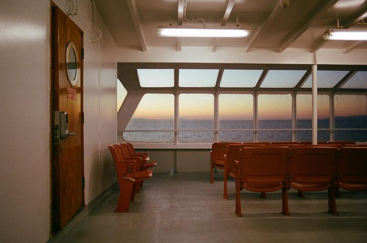 The interior of a ferry with rows of orange seats faces large windows overlooking the ocean at sunset.