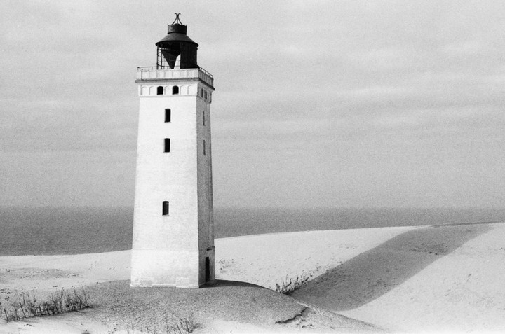 A tall, white lighthouse stands on a sandy dune with the ocean in the background.