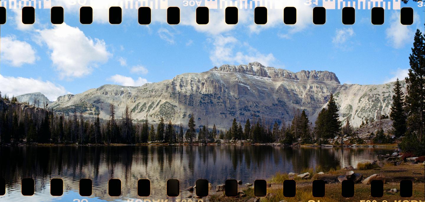 A mountain range with patches of snow rises above a lake surrounded by pine trees under a blue sky with clouds.