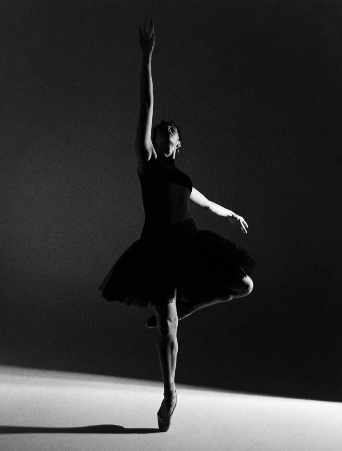 A ballerina in a black tutu and pointe shoes balances on one leg with her arms extended.