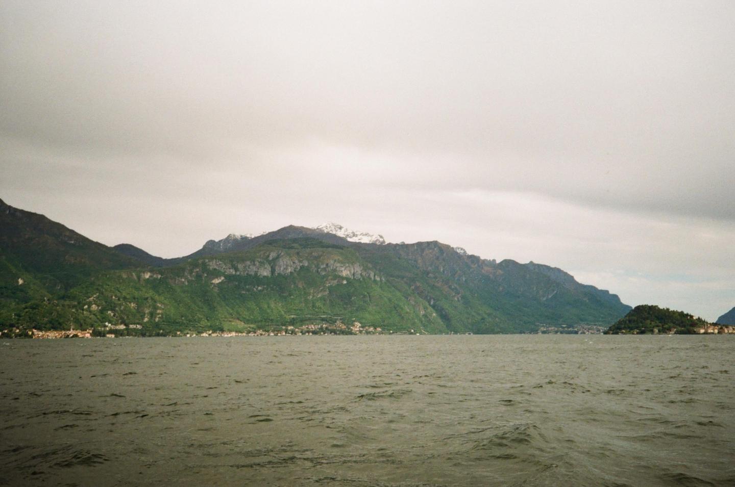 A wide body of water with small waves is in the foreground, with a long, green mountain range and a small town along the shore in the background under a cloudy sky.