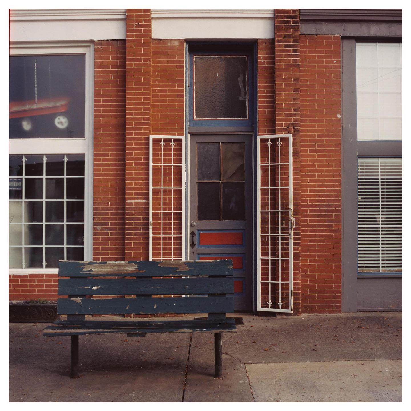 A weathered blue bench sits on a sidewalk in front of a brick building with a door and windows.