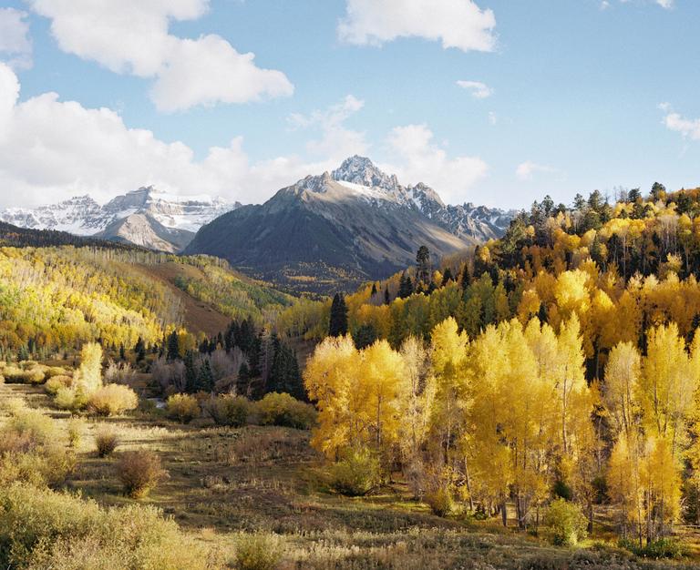 A valley filled with golden trees and green pines is overlooked by snow-capped mountains under a blue sky with white clouds.