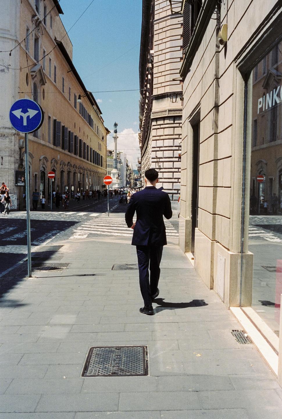 A man in a suit walks away from the camera on a city street with tall buildings and a crosswalk.