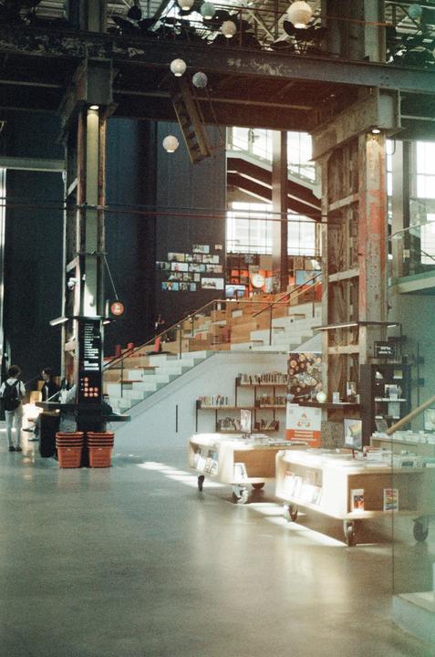 A large, modern library interior features a wide staircase, bookshelves, and people browsing.
