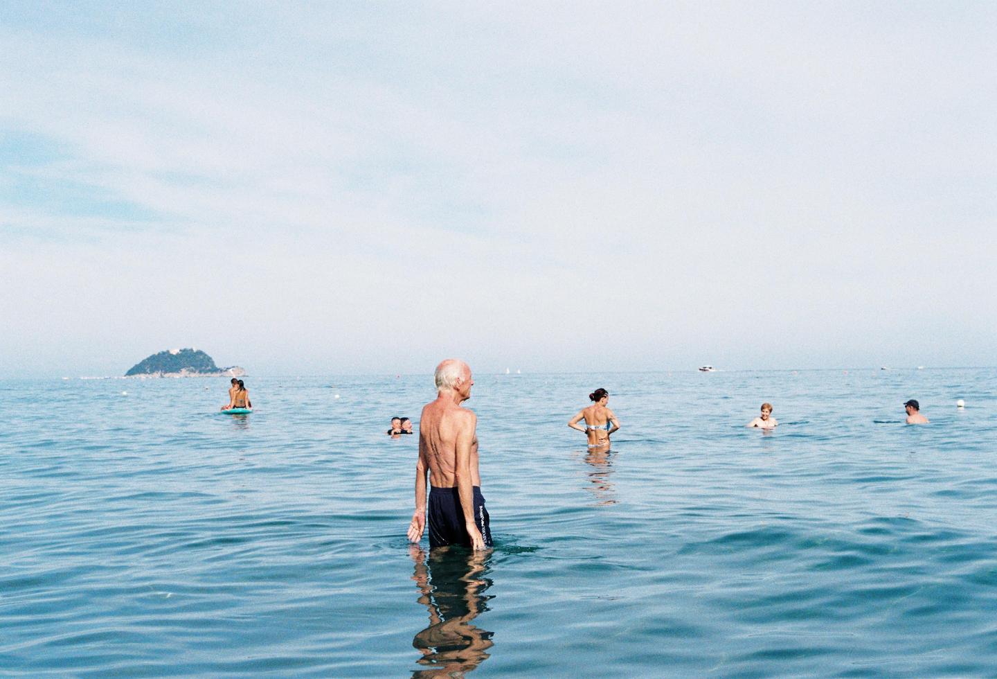 A man stands in the ocean with other people swimming in the background and an island in the distance.