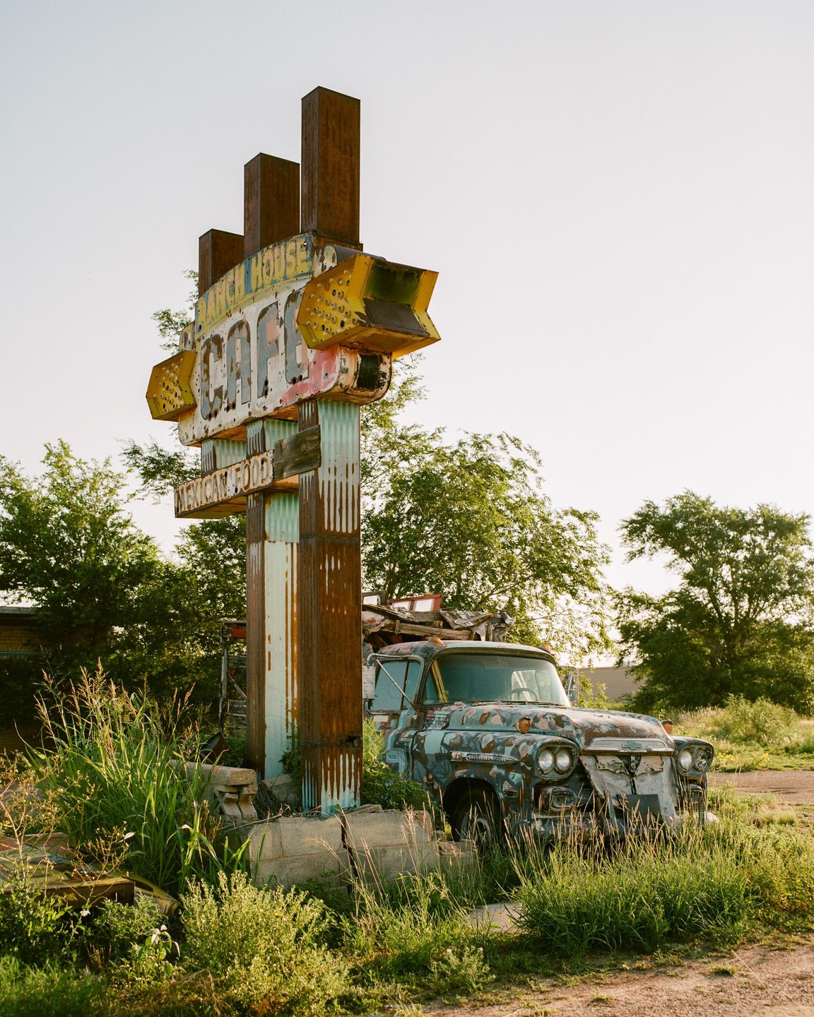 An old, rusty sign for a cafe stands next to a dilapidated truck in an overgrown field.