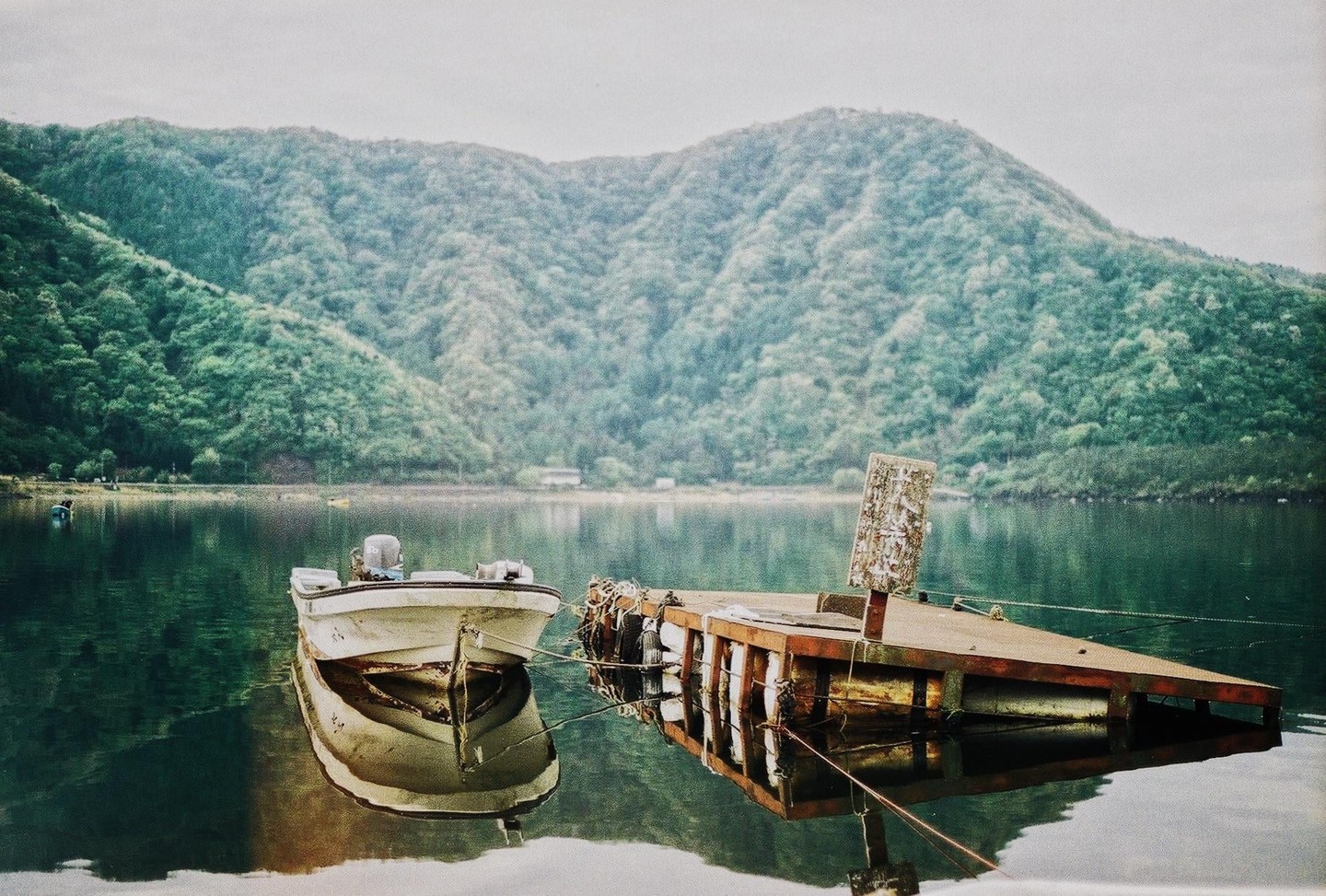 A small boat is moored next to a wooden dock on a calm lake, with a lush green mountain in the background.