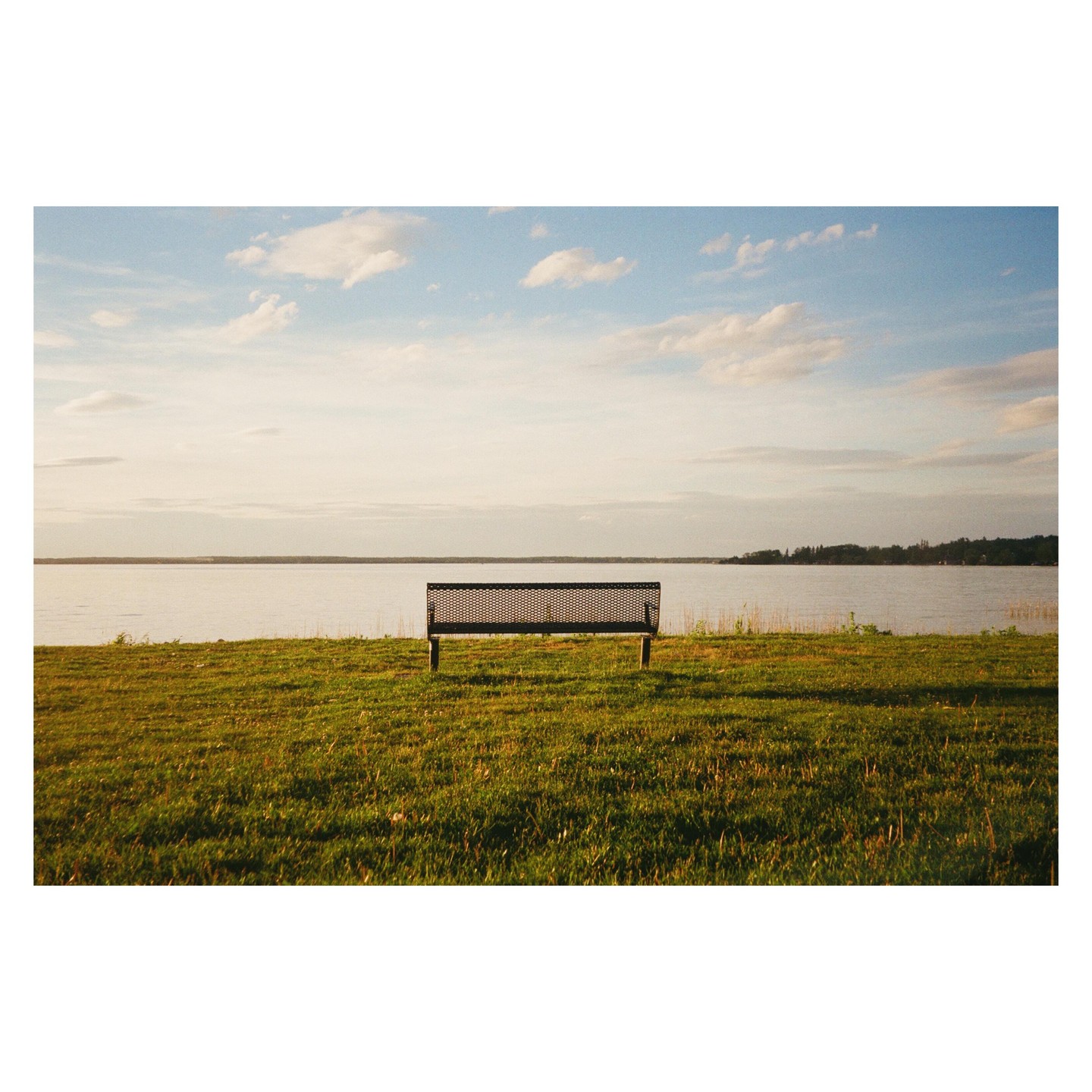 A lone park bench sits on a grassy bank overlooking a wide lake under a partly cloudy sky.