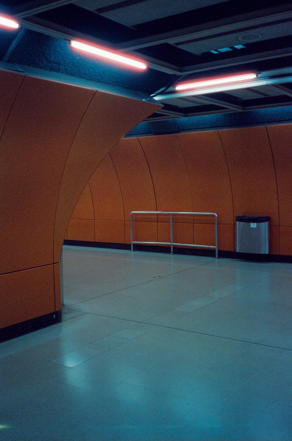 A subway station with orange walls and blue-tinted fluorescent lights has a metal railing and a trash can.