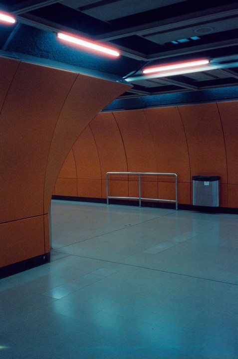 A subway station with orange walls and blue-tinted fluorescent lights has a metal railing and a trash can.