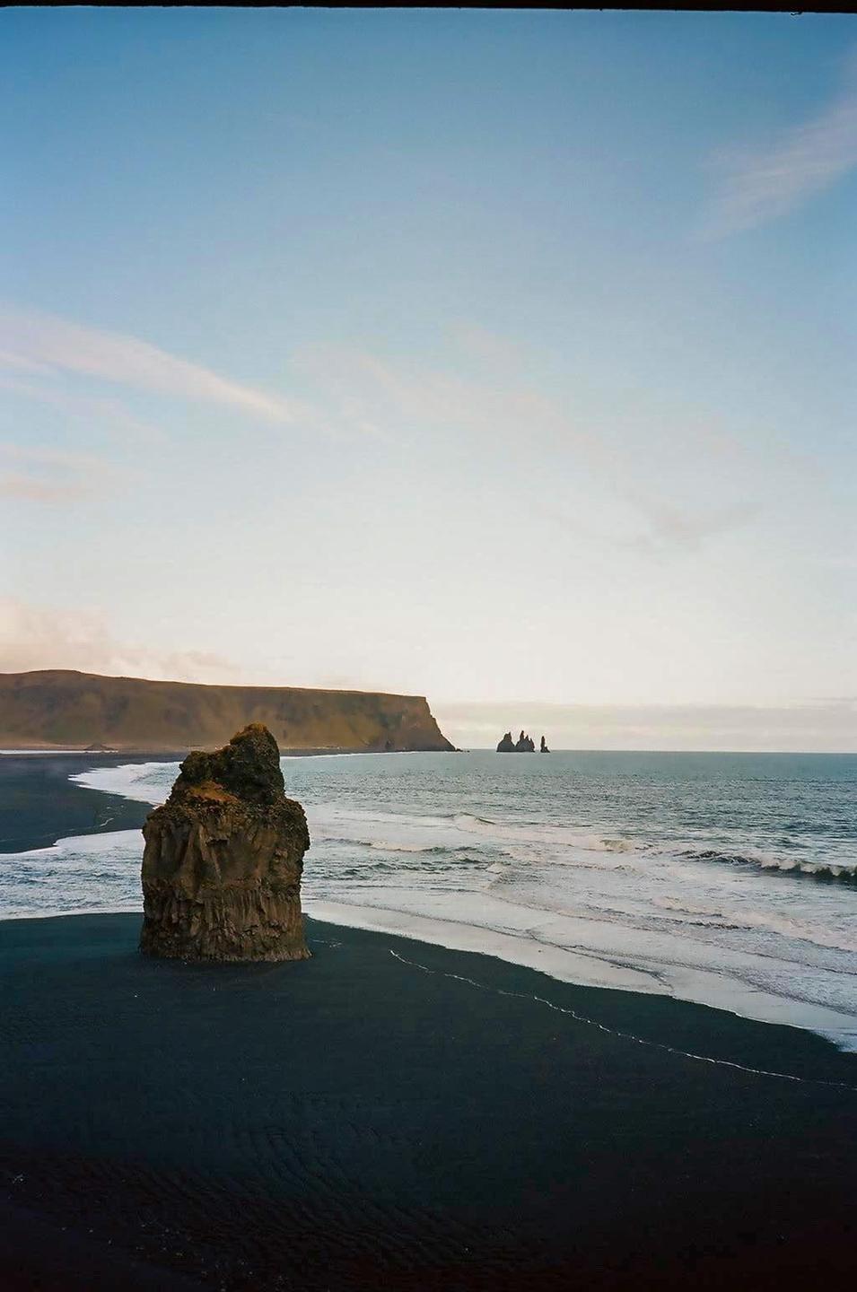 A large rock formation stands on a black sand beach with waves crashing on the shore and cliffs in the distance under a blue sky.