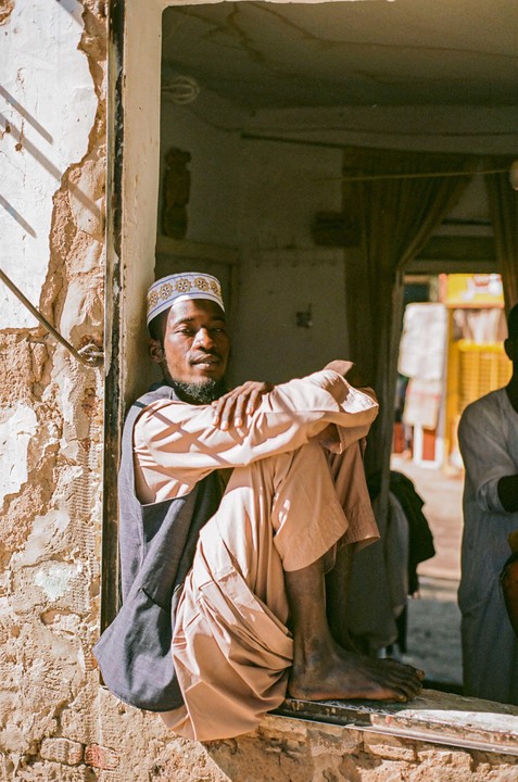A man wearing a kufi and traditional clothing sits in a window frame, looking towards the viewer.
