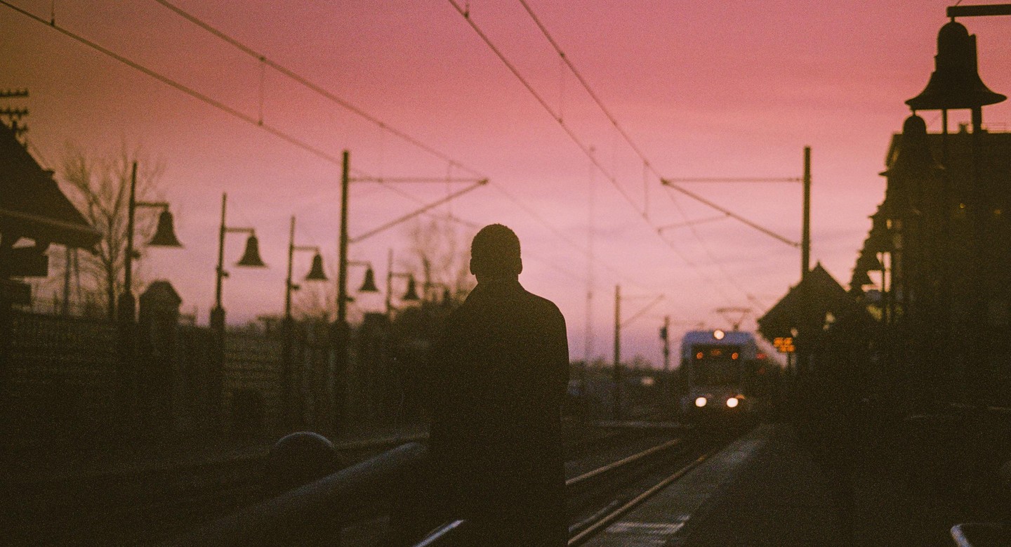 A man stands on a train platform with a train approaching in the distance under a pink sky.