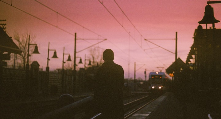 A man stands on a train platform with a train approaching in the distance under a pink sky.