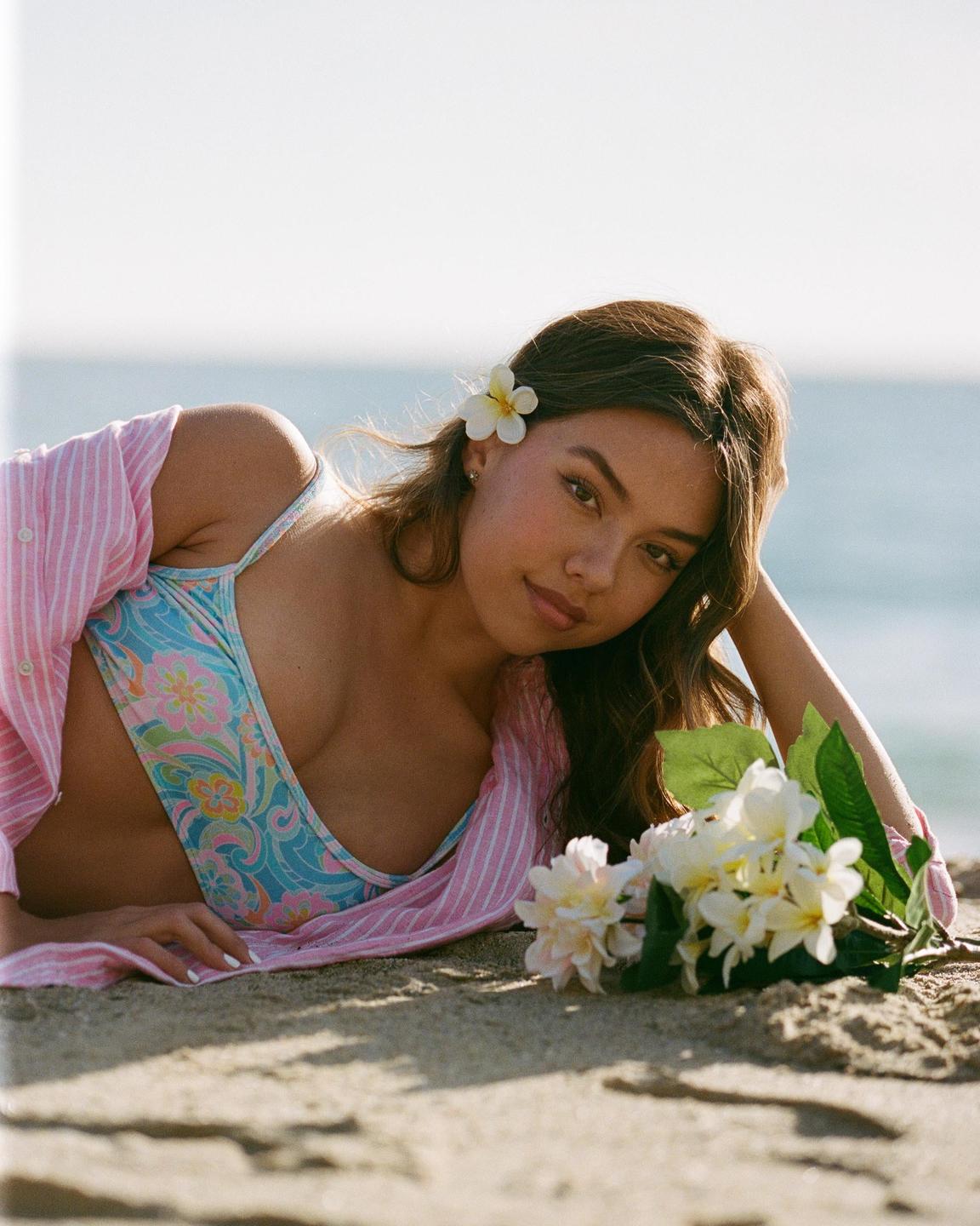 A woman with a flower in her hair is lying on a sandy beach next to a bouquet of flowers.