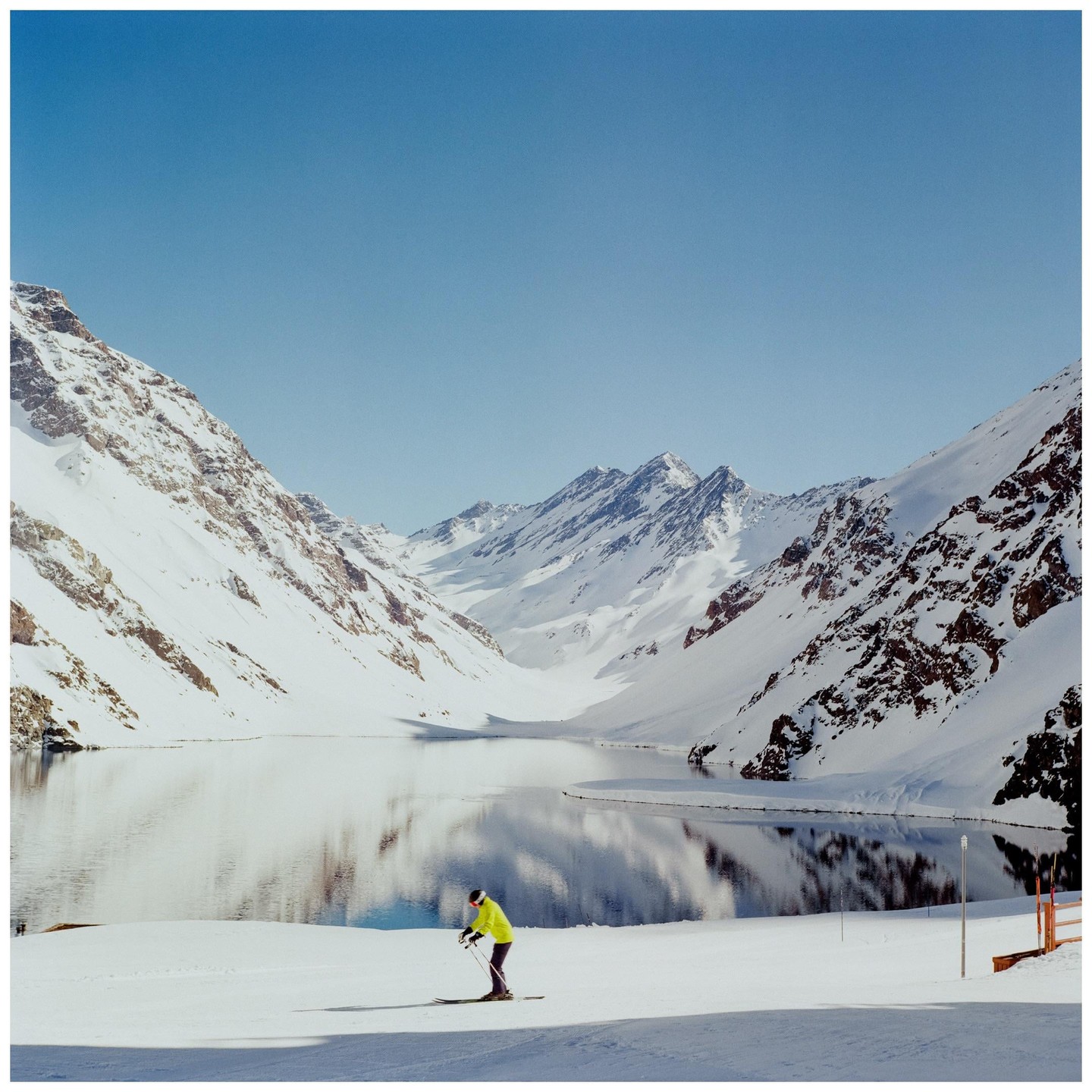 A skier in a yellow jacket stands on a snowy slope next to a lake reflecting snow-covered mountains under a clear sky.
