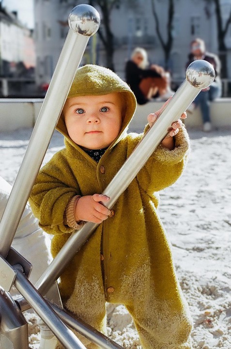A baby in a green hooded suit stands in a sandbox, holding onto metal bars.