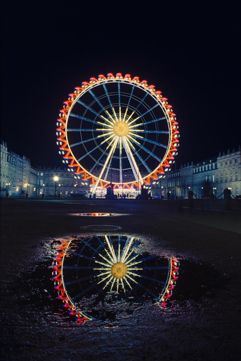 A brightly lit ferris wheel is reflected in a puddle on the ground at night.