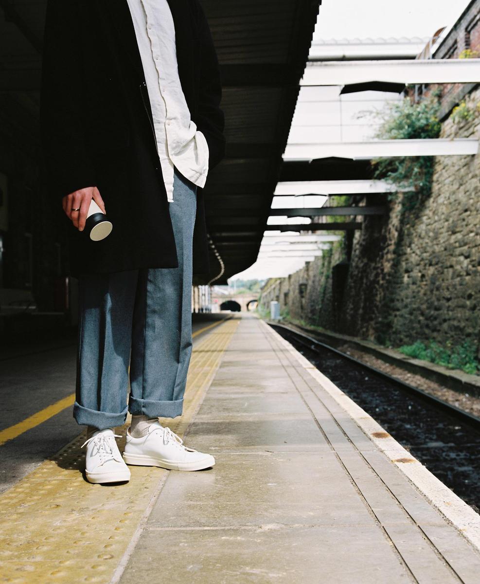 A person stands on a train platform holding a cup, with tracks and a tunnel visible in the distance.