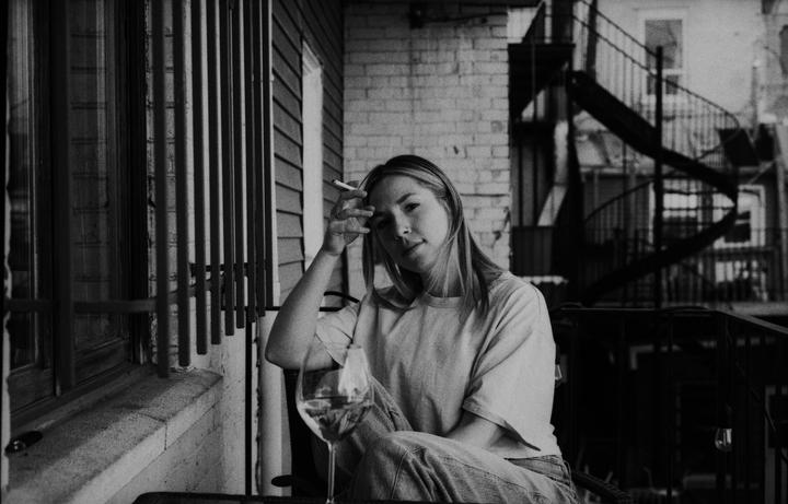 A woman sits on a balcony with a cigarette and a glass of wine.
