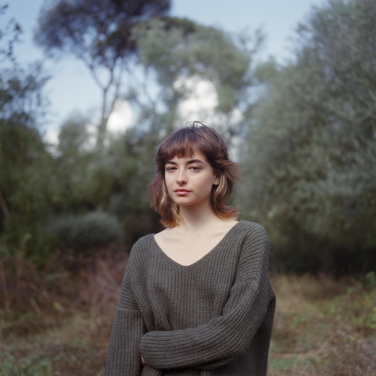 A woman with a bob haircut and bangs wears a green sweater and stands in a forest.