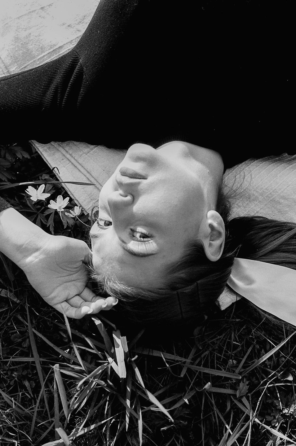A woman lies on her back in the grass with small white flowers nearby.