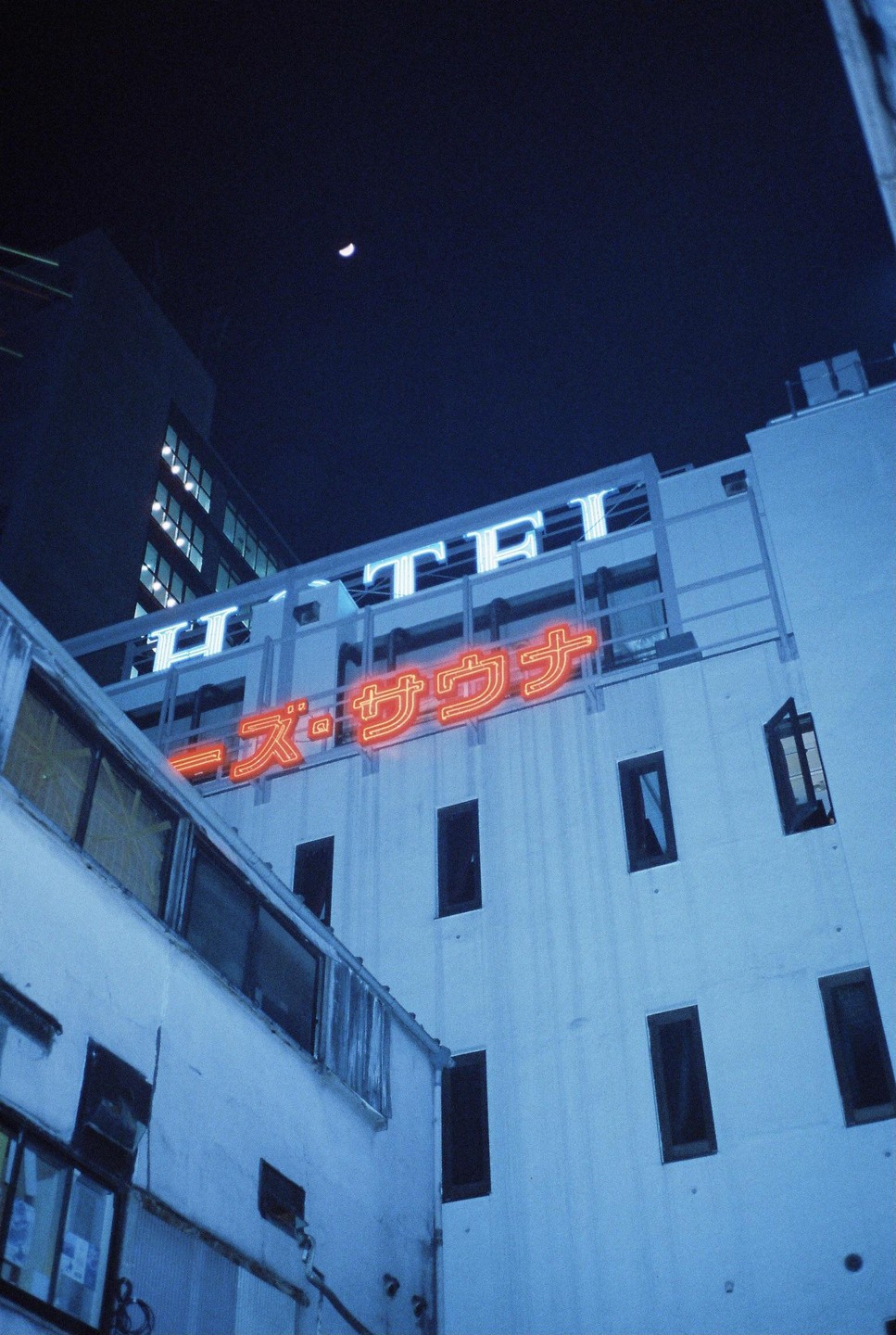 A crescent moon hangs over a building with neon signs for a hotel and a sauna at night.