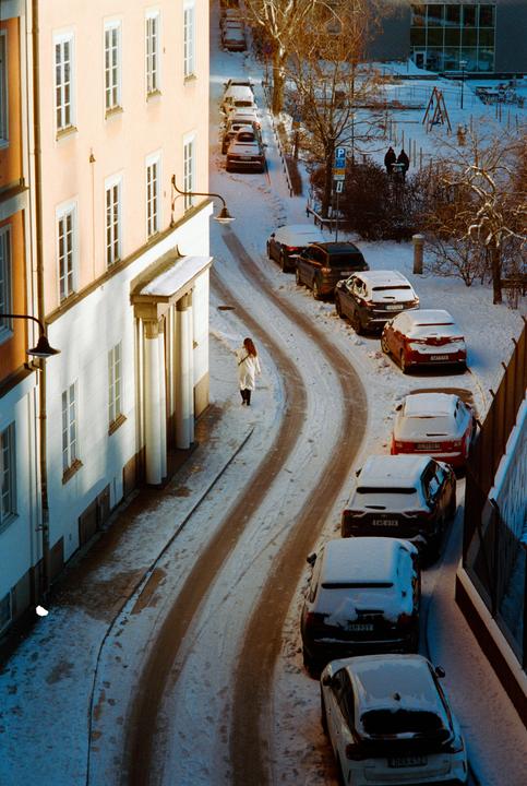 A woman walks along a snow-covered street lined with parked cars and buildings.