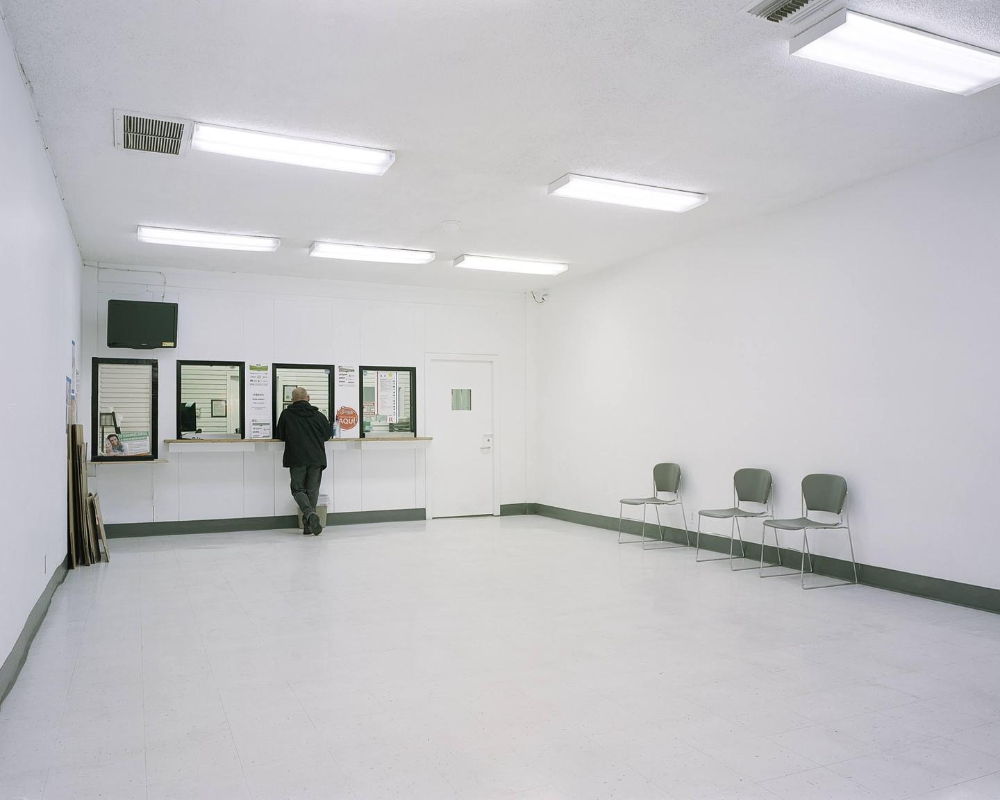A man stands at a service counter in a brightly lit, empty room with several chairs lined against a wall.