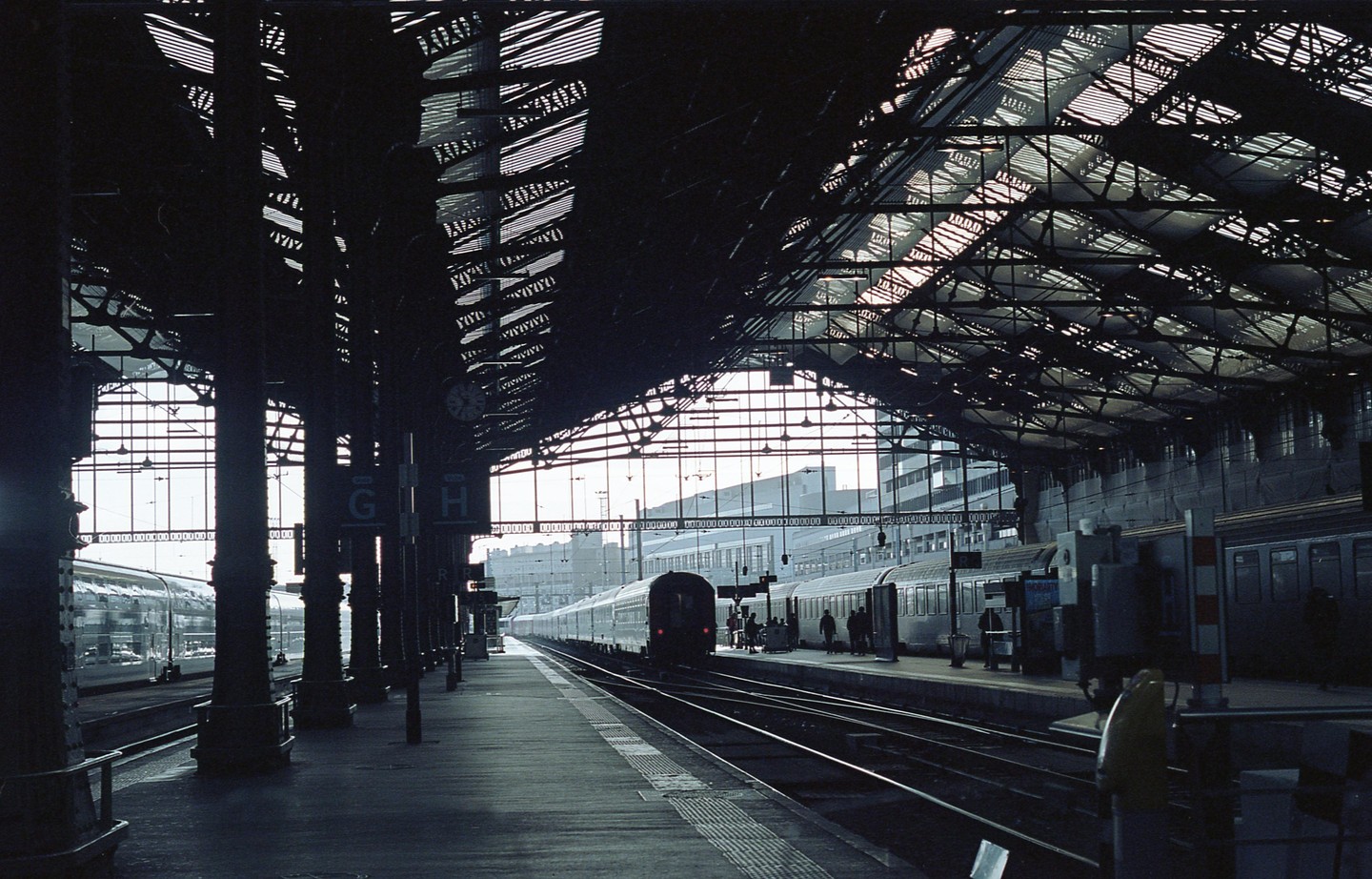 A train station platform is visible with trains on the tracks and people waiting on the platform under a large, arched roof.