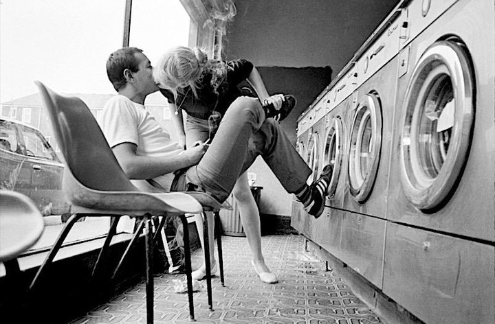 A couple kisses in a laundromat while the man sits on a chair and the woman leans against a washing machine with her leg up.