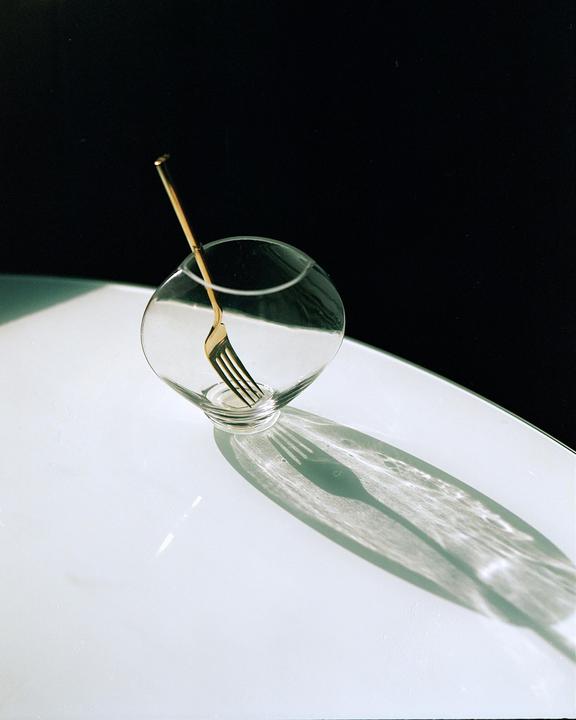 A golden fork is placed inside a clear glass bowl, casting a shadow on a white surface.