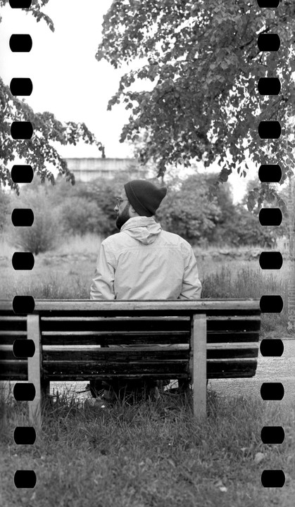 A man wearing a beanie and a jacket sits on a park bench with his back to the viewer, looking towards trees and bushes.