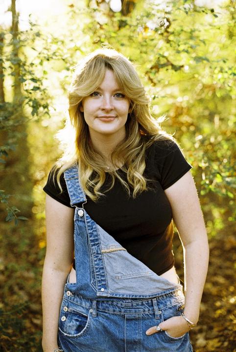 A woman with long hair and denim overalls stands in a forest with sunlight filtering through the trees.