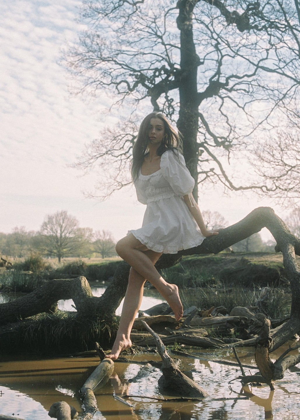 A woman in a white dress sits barefoot on a tree branch over muddy water.