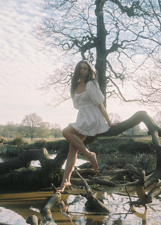 A woman in a white dress sits barefoot on a tree branch over muddy water.