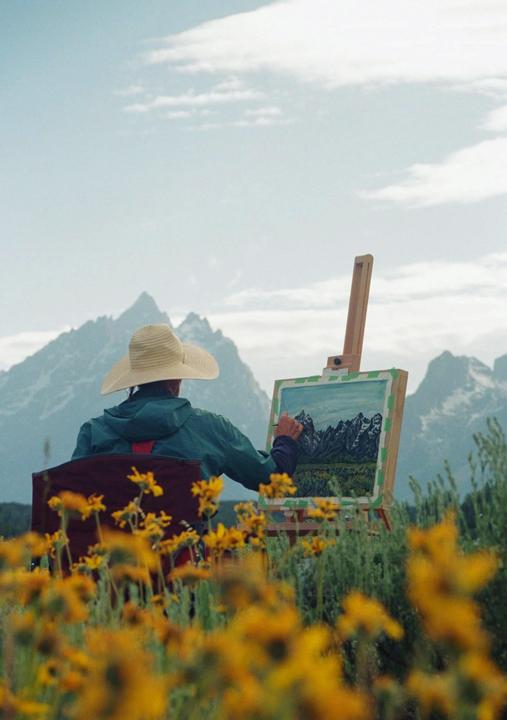 A person wearing a straw hat and a green jacket is painting a mountain landscape on an easel, surrounded by yellow flowers.